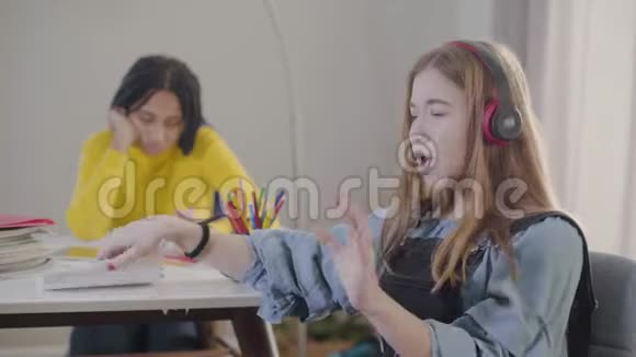戴着耳机的白人少女扔掉笔记本唱歌可爱的青少年作为她的非裔美国朋友休息视频的预览图