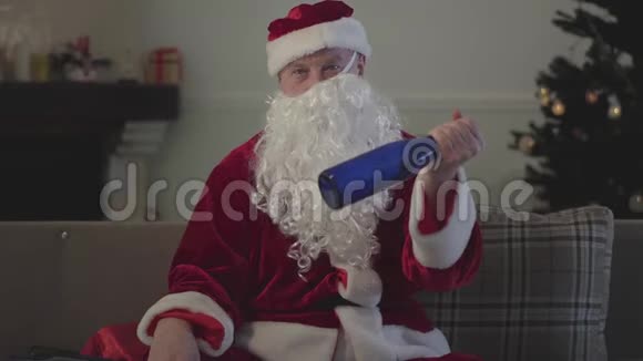 醉汉假胡子歪在圣诞老人的脸上穿着圣诞老人的服装坐在家里的沙发上抱着视频的预览图