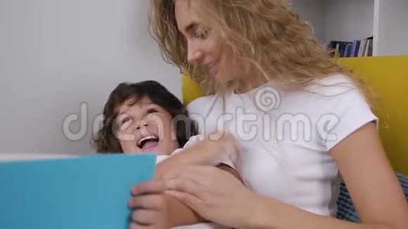 一个漂亮的金发母亲微笑着在看书的时候她那快乐的五岁儿子坐在椅子上挠痒痒视频的预览图