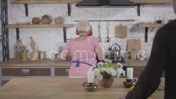 从成熟的白人男人的角度看他的妻子戴着耳机跳舞的金发老太太在厨房做饭视频的预览图