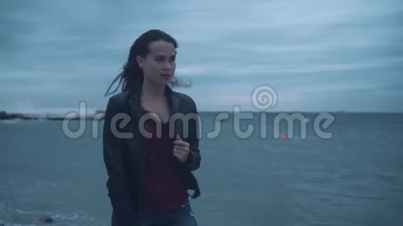 一个穿着自行车夹克的长发冻僵的女孩在暴风雨中站在海滨又阴又冷的一天视频的预览图