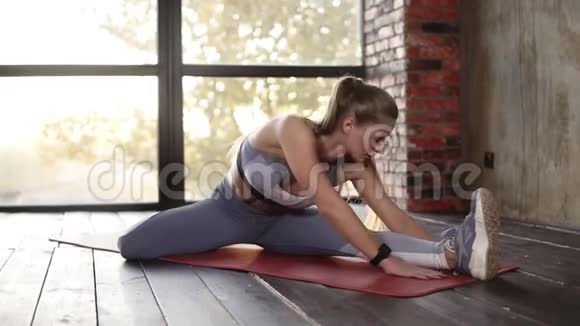 穿灰色运动服和运动鞋的白种人女孩坐在瑜伽垫上一条腿向前弯着身子坐着视频的预览图
