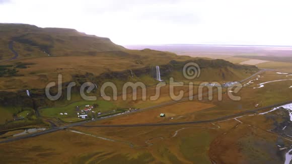 冰岛广阔的空中山景美丽的绿色平原和一个小村庄附近的瀑布一个大山的无人机视频的预览图