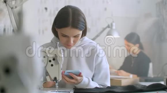 女裁缝看着工作场所的智能手机屏幕一位同事在缝纫机上工作和缝纫A视频的预览图