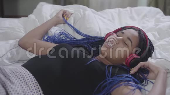 戴耳机的十几岁非裔美国女孩的特写镜头她躺在白色的床上闭着眼睛做手势动着头视频的预览图