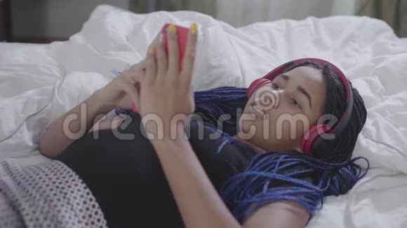非洲裔美国少女戴耳机躺在软床上用智能手机打开音乐和关闭的肖像视频的预览图