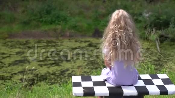 一个金发碧眼的小女孩坐在公园池塘边的格子长凳上视频的预览图