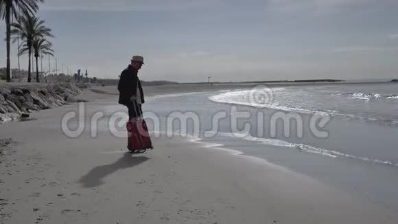 一个戴着帽子带着红色手提箱的人来到海边走近海浪看着风景视频的预览图