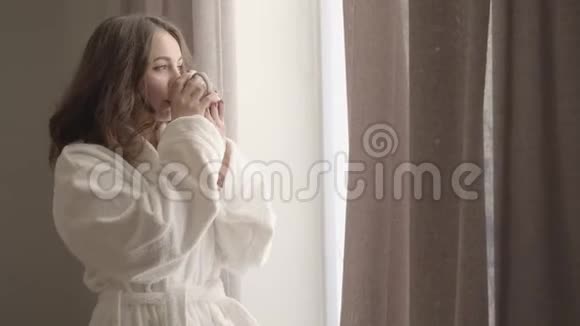 穿着白色柔软浴袍的迷人年轻女士在家享受阳光明媚的早晨的肖像可爱的白种人黑发女人视频的预览图