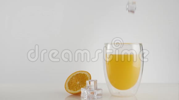 用冰块装饰在白色桌子上的玻璃杯中冰块随着飞溅和气泡落入新鲜的橙汁或菠萝汁中视频的预览图