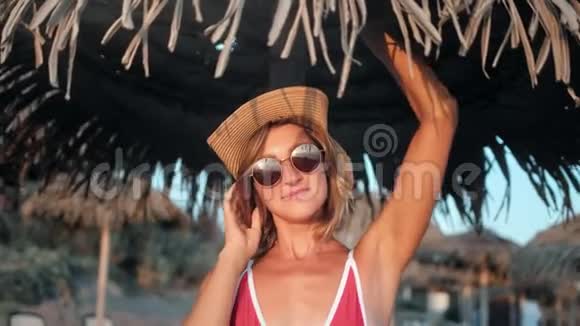 穿着比基尼的快乐时尚旅行女孩在沙滩上与棕伞合影视频的预览图