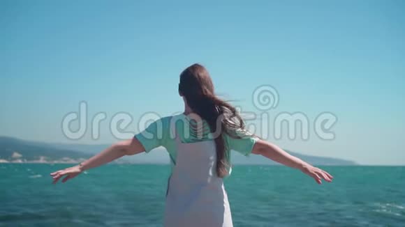 一个穿着蓝色T恤和工作服的女孩站在海边女孩举起双手显示出自由的迹象视频的预览图