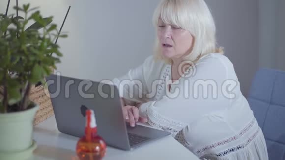 侧面观看漂亮的金发白种人成熟的女人坐在家里的笔记本电脑站在她旁边的绿色植物视频的预览图