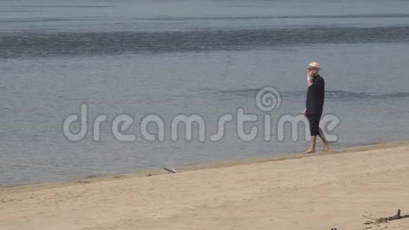 戴着太阳镜戴着帽子的人正慢慢地沿着海滩上的沙子沿着水走在打电话视频的预览图