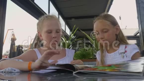 两个女孩在户外夏季咖啡馆寻找菜单年轻女孩青少年在餐厅菜单上交谈和选择食物视频的预览图