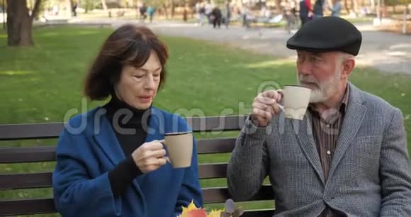 幸福的老家庭正在舒适的城市公园里的长凳上休息秋天老人夫妻在公园里喝茶视频的预览图