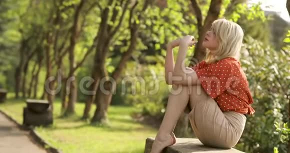 年轻迷人的女人一头华丽的金色头发正在美丽的夏季公园里休息她坐在长凳上视频的预览图