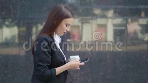 一个留着长发的女人穿着漂亮的休闲服手里拿着外卖咖啡杯在街上走着一边发短信视频的预览图