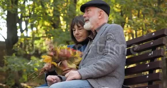 快乐的老年家庭在秋天城市公园的新鲜空气中悠闲地坐在长凳上妻子抚摸她视频的预览图