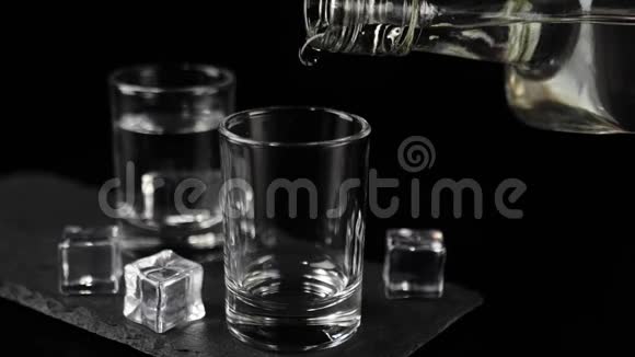 用手将伏特加龙舌兰酒或清酒从扁平的瓶子中倒入装有冰块的黑色木板上的空杯中视频的预览图