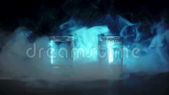 在黑色背景下冰块从两杯伏特加龙舌兰酒或清酒中掉到一杯蓝烟云雾视频的预览图