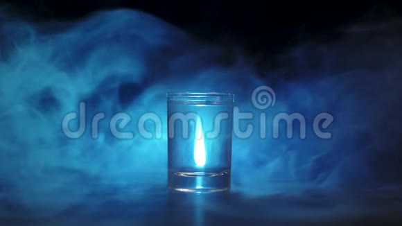 冰块在黑色背景下落入伏特加龙舌兰酒或清酒的杯中蓝色烟雾云雾或蓝色蒸汽视频的预览图