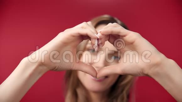 近距离微笑的年轻女子在红色摄影棚背景下用手摆姿势做心形标志视频的预览图