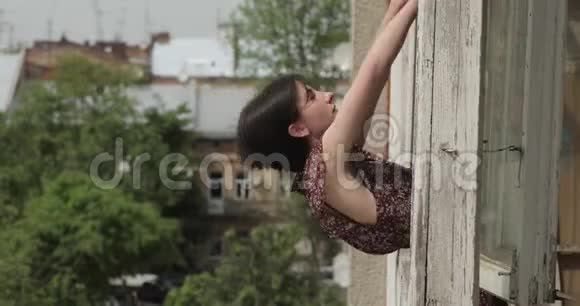 年轻的黑发女人躺在窗台上的窗户上背景中的老建筑和绿树4K视频视频的预览图