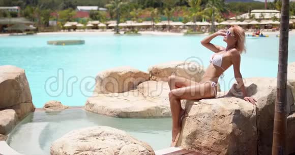 穿着白色泳装坐在池边石头上享受热天的优雅性感女人视频的预览图