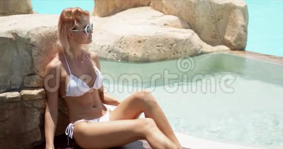 穿着白色泳装的迷人女人靠在游泳池附近的石头上对着镜头微笑视频的预览图