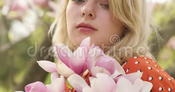 一位年轻迷人的女人的肖像她有一头华丽的金色头发在粉红色花瓣的花树之间可爱的金发美女视频的预览图