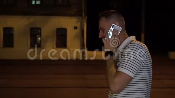 留胡子的男人穿着白色和黑色的条纹T恤晚上在街上散步用手机或手机聊天视频的预览图