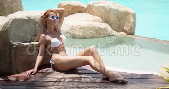穿着白色泳装享受热天坐在游泳池旁的木制露台上的宁静性感女人视频的预览图