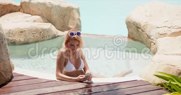 游泳池中的美女在阳光下浏览智能手机视频的预览图