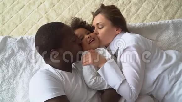 年轻幸福的多民族家庭的母亲非洲父亲和小女儿穿着白色衣服躺在床上视频的预览图