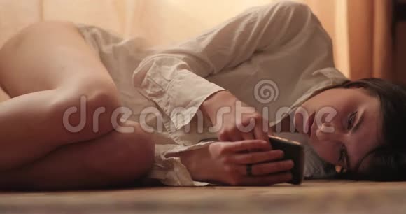 一个漂亮的女人躺在木地板上的毯子上在阁楼的公寓里通过手机在社交网络聊天视频的预览图