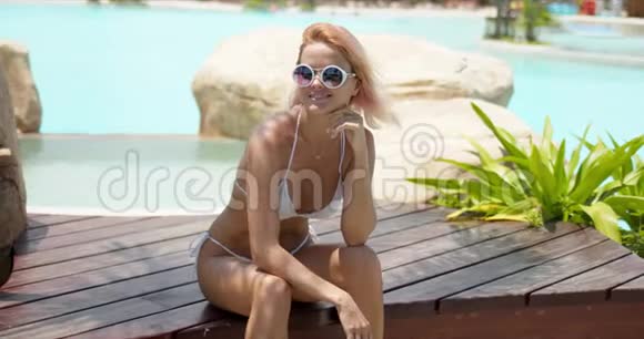 穿着白色泳装的开朗女人坐在游泳池旁的木制露台上视频的预览图