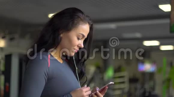 戴着耳机的年轻女孩在运动自行车上使用智能手机视频的预览图