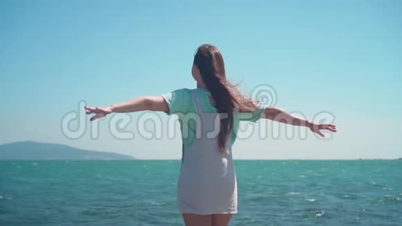 一个穿着蓝色T恤和工作服的女孩站在海边女孩举起双手显示出自由的迹象视频的预览图