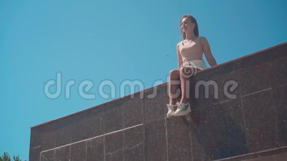 一个穿白色短裤和T恤的女孩沿着城市长廊一个女孩坐在花岗岩栅栏上视频的预览图
