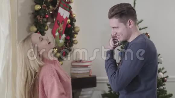 侧面观看快乐的白种人夫妇在家的圣诞树前欢笑和拥抱积极的家庭开支视频的预览图