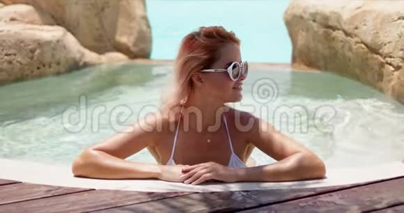 戴墨镜的女人在凉爽的游泳池里躺着喝着水晶水视频的预览图