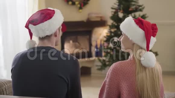 年轻快乐的白人夫妇坐在圣诞树前的后景戴着红色圣诞帽的女人视频的预览图