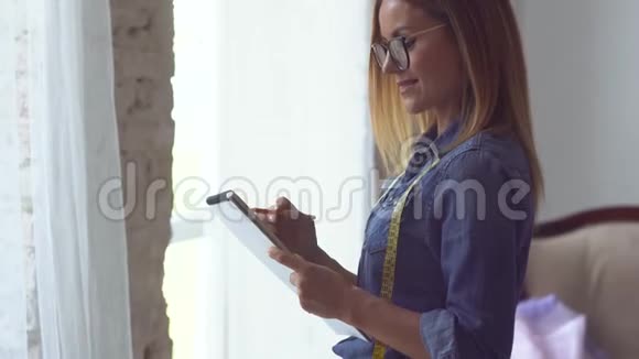 美丽的金发女裁缝站在窗边脖子上缠着一米长的胶带手里拿着一个笔记本做着视频的预览图