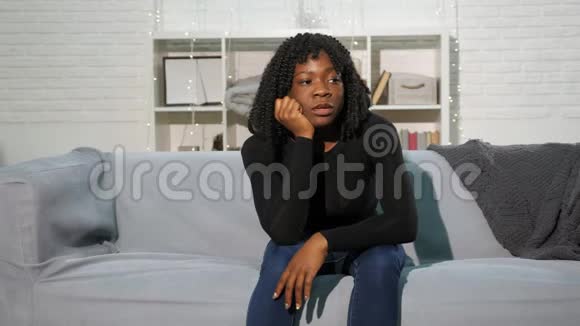 无聊的黑人女孩坐在灰色的沙发上靠在白色的墙上慢慢地视频的预览图