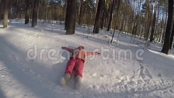 一个穿着桃色夹克和粉红色暖裤的女孩躺在雪堆里手举起来在毡靴的腿上清澈的冰霜视频的预览图