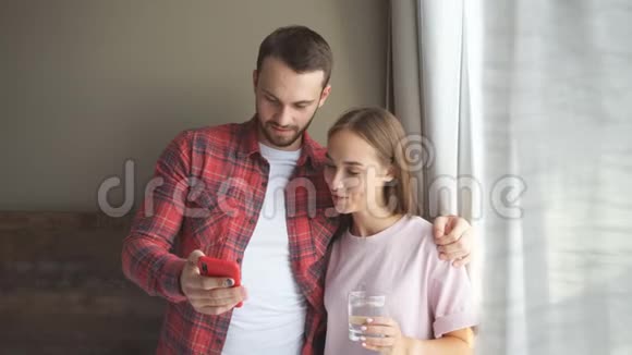 早晨一对快乐的年轻夫妇站在明亮的窗户旁在家看有趣的视频视频的预览图
