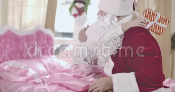 无法辨认的金发白种人女孩坐在粉红色的床上拥抱着穿着圣诞老人服装的男人快乐的孩子感谢视频的预览图
