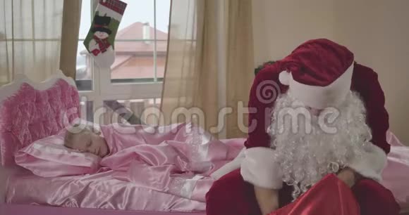 白种人圣诞老人坐在粉红色的床上带着红色的礼品袋金发女孩睡在背景上的肖像视频的预览图