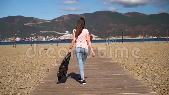 年轻苗条的女孩或年轻女子穿着粉红色的t恤走在木路上手里拿着夹克慢动作视频的预览图